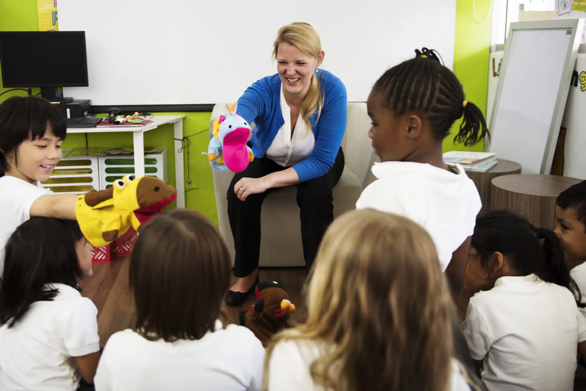 A woman sits in an educational classroom with children who are holding hand puppets. The children are seated on the floor and facing the woman, who is smiling and holding two colorful puppets.