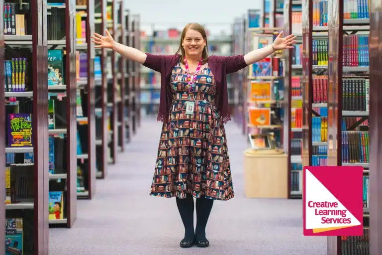 A bookworm stands in the aisle of a library with arms outstretched, wearing a dress printed with books. Shelves of colorful books line both sides. The "Creative Learning Services" logo is visible.