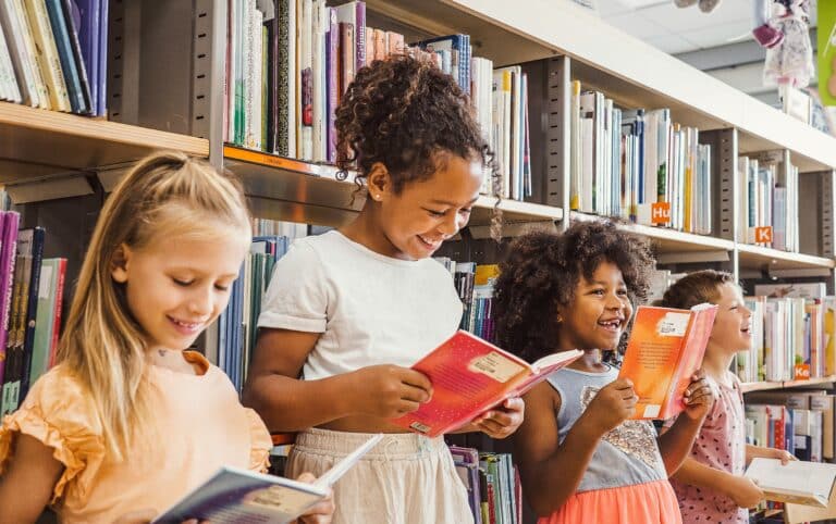 Four children stand in a school library aisle, smiling and reading books in front of shelves filled with colorful books.